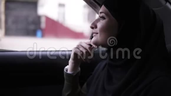 开朗迷人的女人戴着黑色头巾头巾在车里看着窗户微笑的女人享受她的路风视频的预览图
