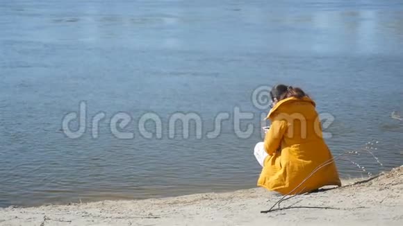带着智能手机坐在河岸上的女孩晴朗的秋天天气慢动作视频的预览图