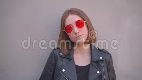 年轻可爱的白种人女孩穿着皮夹克和戴着红色太阳镜在面前无聊地摆姿势的特写照片视频的预览图