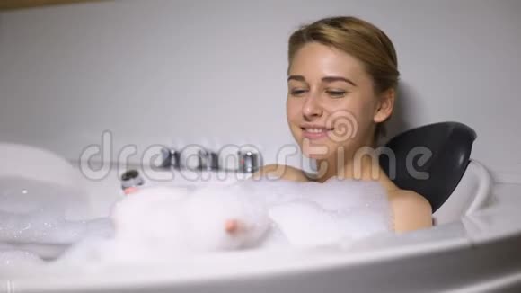 有趣的女性躺在浴缸里把泡沫泡泡吹进相机孩子气的心情视频的预览图