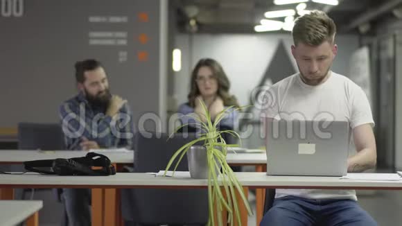 这位穿着白色t恤手提电脑的年轻人坐在现代办公室的桌子旁视频的预览图