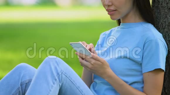 年轻的微笑女性在智能手机上聊天在阳光明媚的公园里放松视频的预览图