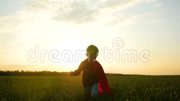 一个穿着超级英雄服装的快乐的孩子穿过绿草在夕阳的背景下跑到镜头前视频的预览图