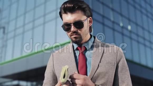 穿着时髦西装戴着墨镜拿着一卷钞票站在办公室附近的街道上的富人画像视频的预览图