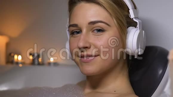 戴耳机的快乐女人用泡沫在浴室里放松微笑着进入相机休息视频的预览图