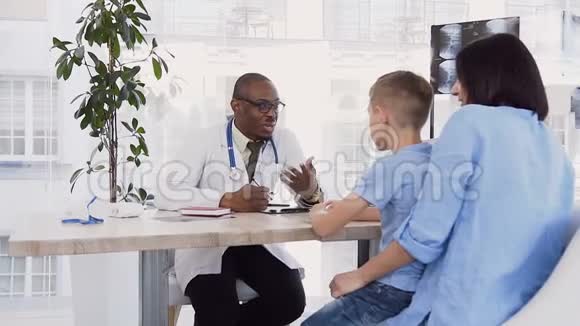 非裔美国医生在诊所里和漂亮的白种人母亲和儿子谈话视频的预览图