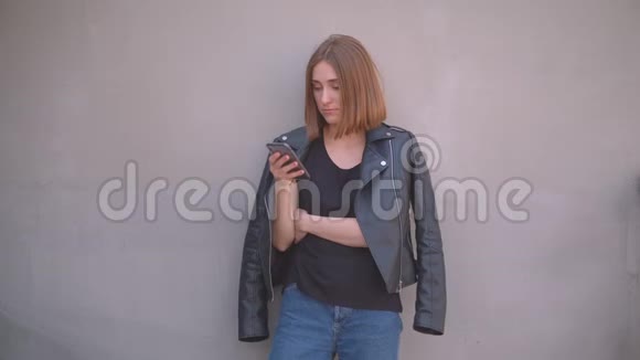一个年轻可爱的白种人女孩穿着皮夹克在室外镜头前用手机特写照片视频的预览图