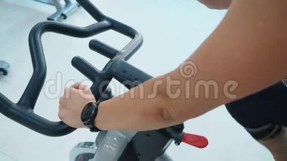 在体育俱乐部的自行车课上带健身手表的女性手拿着室内自行车的车把斯利姆妇女培训视频的预览图