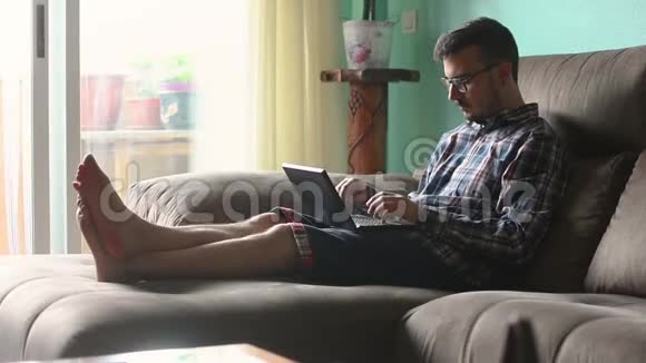 年轻人正在他家沙发上看着他的平板电脑和智能手机视频的预览图