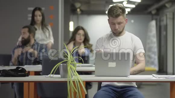 这位穿着白色t恤手提电脑的年轻人坐在现代办公室的桌子旁视频的预览图