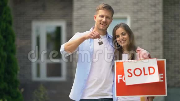 幸福的一对夫妻微笑着手里拿着房子的钥匙站在出售的招牌附近视频的预览图