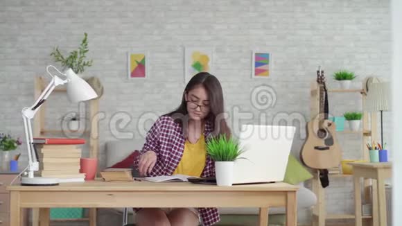 漂亮的亚洲女孩戴着眼镜留着长发在家里的客厅里手提电脑和课本在学习或视频的预览图