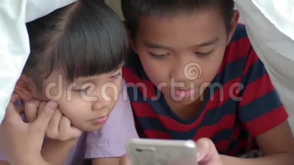孩子们在床上的毯子下面使用智能手机或平板电脑视频的预览图