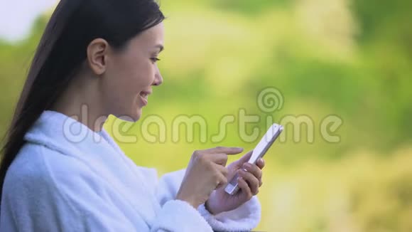 穿着白色浴袍滚动智能手机照片休闲度假的快乐女人视频的预览图