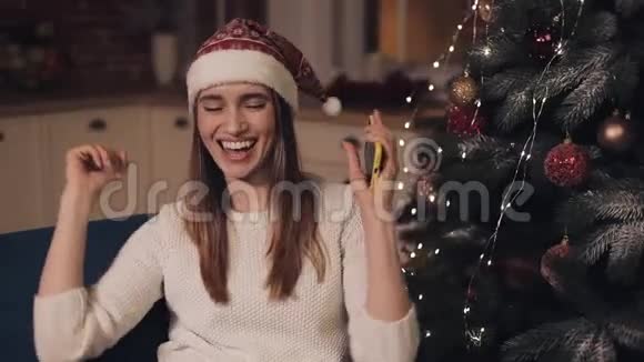 戴着圣诞帽的年轻漂亮女孩手里拿着智能手机坐在沙发上玩得很开心视频的预览图