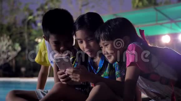 孩子们晚上使用智能手机在游泳池附近放松视频的预览图