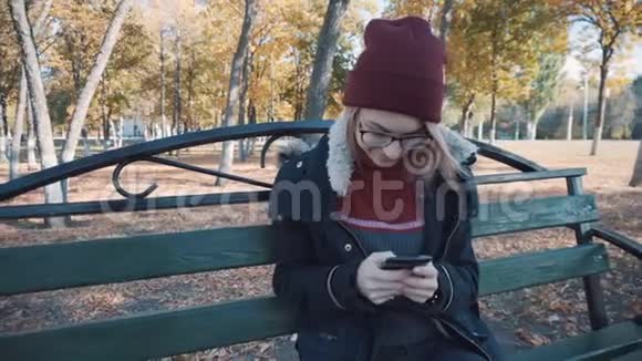 带着手机坐在公园的长凳上的女孩穿着夹克和帽子的女孩视频的预览图