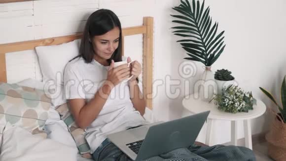 年轻女子坐在床上喝茶手提电脑看电影慢动作视频的预览图
