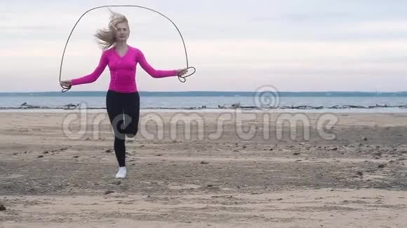 穿白色运动鞋的女孩黑色紧身裤粉红色的t恤在海滩上跳绳健身在大自然夏季阴天在海滩上视频的预览图