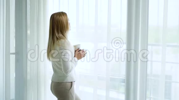 放大漂亮的金发女人白天在大窗帘窗前用杯子做梦视频的预览图