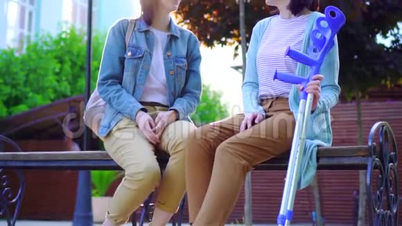 一位拄着拐杖的年轻女子和一位朋友坐在公园的长凳上阳光明媚的一天视频的预览图