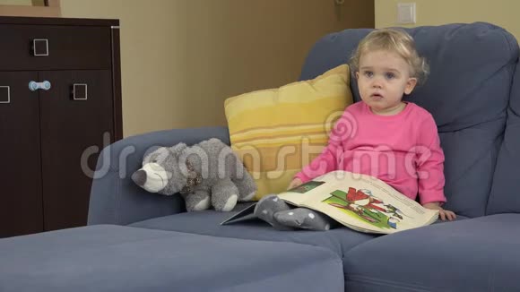 一个好奇的小女孩在看图片书和玩具熊坐在蓝色的沙发上视频的预览图