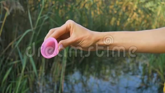 靠近女人手与粉红色硅胶月经杯站在绿色自然背景与湖泊零废物视频的预览图