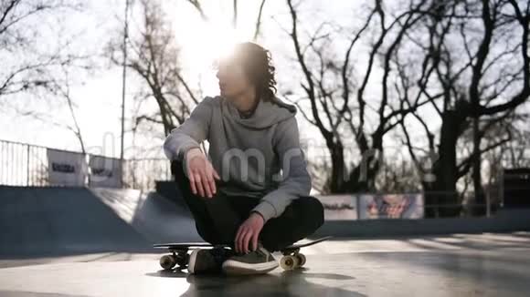 穿着浅灰色连帽衫的溜冰者坐在他的木板上在溜冰公园外面看上去很体贴强烈的阳光视频的预览图