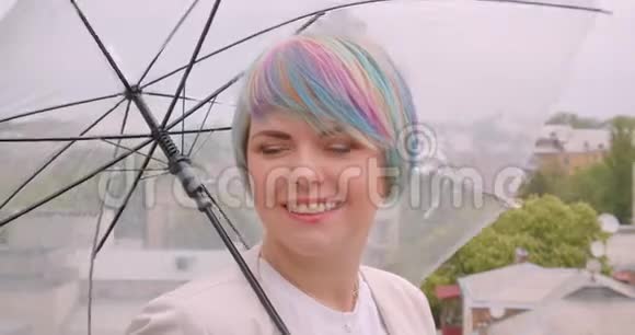 年轻漂亮的白种人女性的特写照片她染着头发拿着雨伞看着室外的摄像机视频的预览图