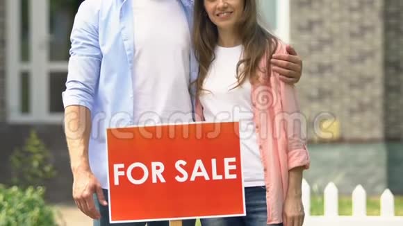 一对幸福的年轻夫妇在出售时站在房子外面的标志微笑相机视频的预览图