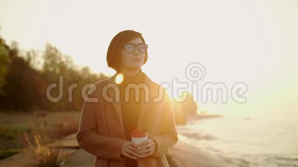 日落时穿着外套戴着眼镜的亚洲女人在海滩上行走的慢动作视频的预览图