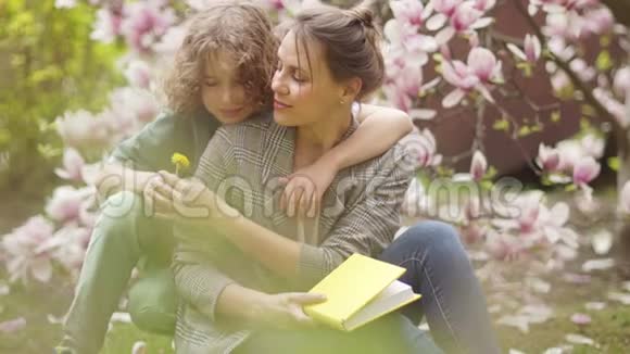 可爱的小学生在盛开的玉兰花的背景下拥抱他的妈妈春天的野餐幸福的家庭视频的预览图