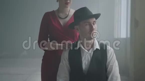 英俊自信的黑手党老板戴着帽子和背心坐在一座废弃的大楼里穿红色裙子的优雅女人视频的预览图