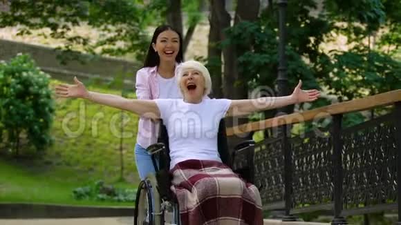 公园里的微笑志愿者和坐在轮椅上的受启发的老妇人举手视频的预览图