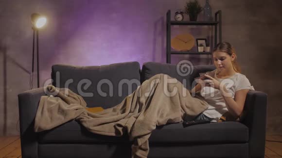 年轻漂亮的女孩躺在沙发上在电话里写信息视频的预览图