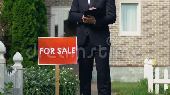 微笑男经纪人西装站在房子外的销售标志附近视频的预览图