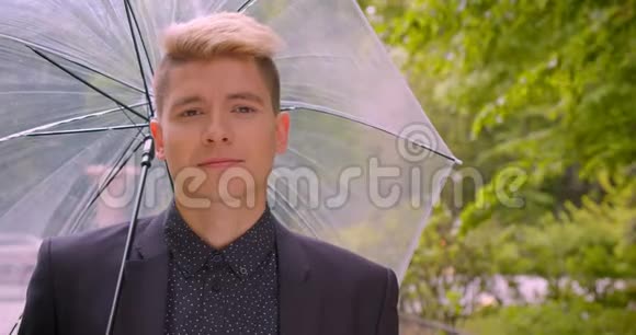 一位年轻迷人的白种人金发男子手持一把伞笑着愉快地看着镜头的特写镜头视频的预览图