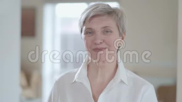 一幅漂亮的穿着白色衬衫的高级女人的肖像在镜头里看着在室内靠近时露出了愉快的微笑背景视频的预览图