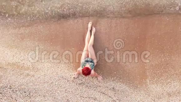 顶景年轻女子穿着泳装坐在沙滩上双腿抚摸着海浪视频的预览图