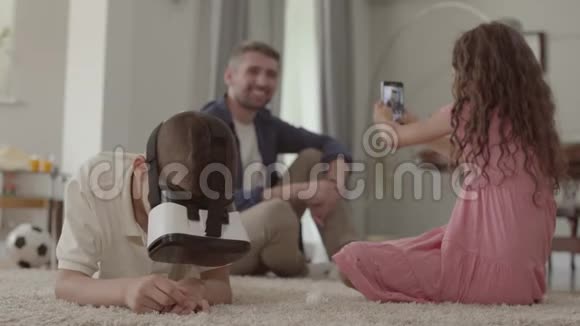 男孩和女孩躺在毛茸茸的地毯上哥哥用虚拟现实眼镜姐姐在拍照片视频的预览图