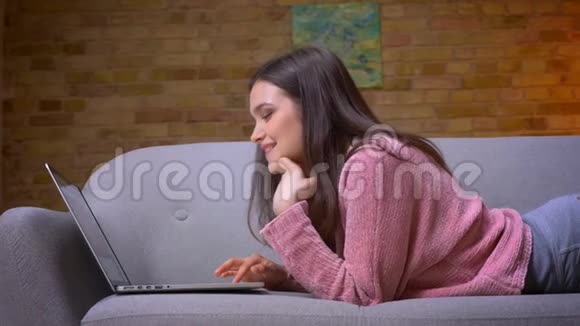 一位年轻漂亮的黑发白种人少女使用笔记本电脑并微笑地躺在沙发上的特写镜头视频的预览图