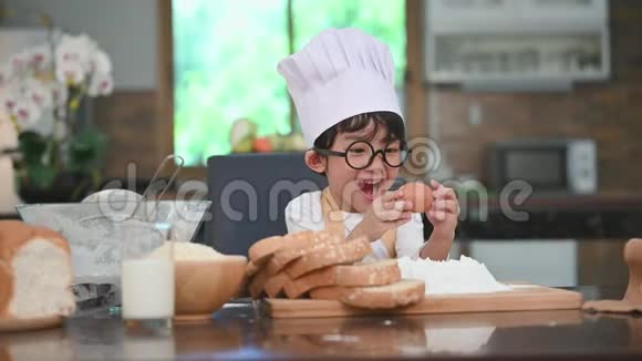 可爱的亚洲小男孩带着眼镜厨师帽和围裙带着鸡蛋第一次体验到家中的烘焙面包店视频的预览图