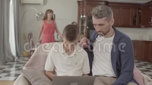 父亲和儿子一起坐在扶手椅上拿着笔记本电脑美丽的母女来到他们身边视频的预览图
