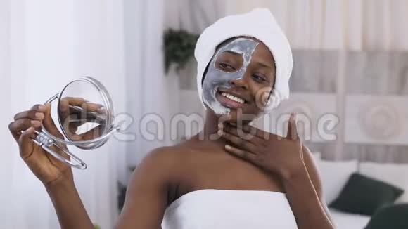 年轻微笑的非洲女人的肖像头上戴着白色毛巾半张脸上戴着粘土面具看着视频的预览图
