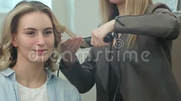 理发师正在为坐在一个微笑的女人面前的卷发熨斗做发型卷发视频的预览图