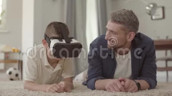 肖像英俊的父亲和儿子躺在毛茸茸的地毯上用虚拟现实眼镜快乐友好视频的预览图