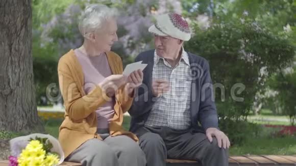 带着一束黄花的老妇人和一位老人坐在绿色美丽的公园里的长凳上给他看视频的预览图