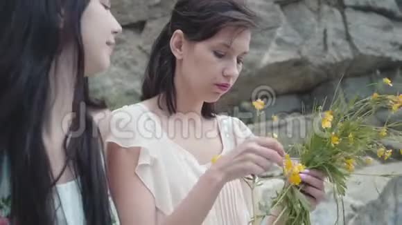 两个美丽的女人黑色的头发编织花冠靠近巨大的石头与绿色的植物可爱的女友们视频的预览图