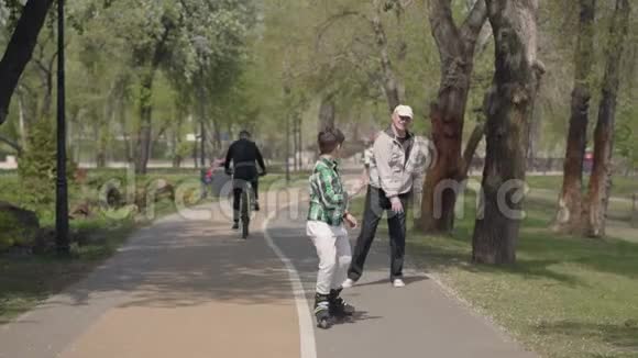 可爱的男孩穿着鲜艳的衣服在公园里滑旱冰他的祖父走在附近户外休闲孩子玩得很开心视频的预览图