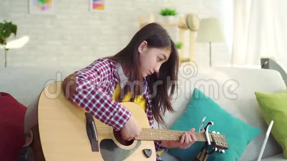 美丽的亚洲女孩留着长发在现代家庭的客厅里在网上观看吉他训练视频的预览图